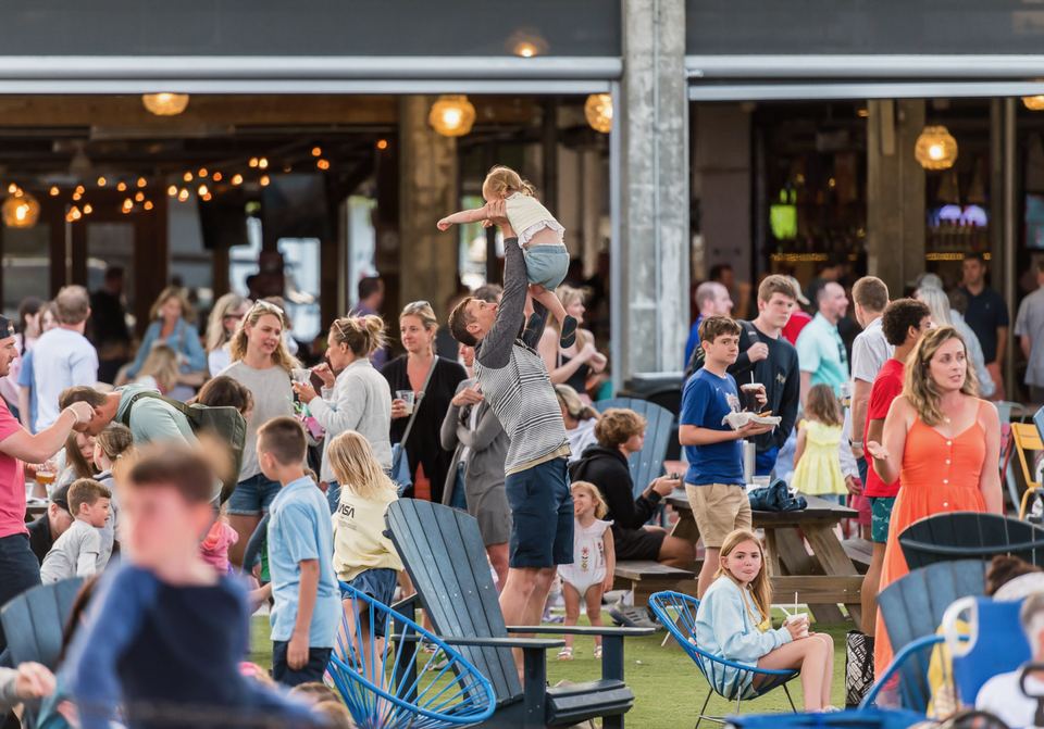 A dad lifting a toddler overhead on the lawn, families and friends all around at dusk